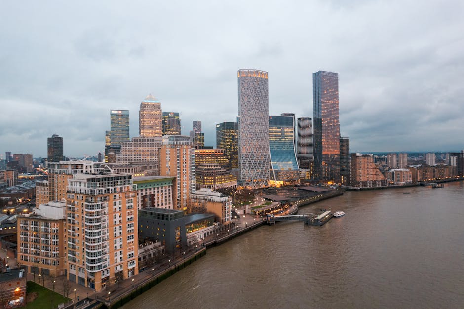 A panoramic view of a modern city skyline with numerous high-rise office buildings and glass skyscrapers, set against a bright blue sky with scattered clouds. In the foreground, there is a lush green 