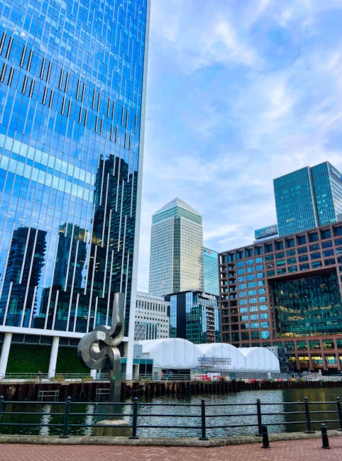 Nighttime view of the Canary Wharf skyline reflected on the calm river, with illuminated modern skyscrapers and office buildings featuring glass and steel facades. The city lights create a shimmering 